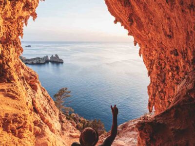 Hiker standing at the edge of a cave balcony, gazing out over sweeping views of forested cliffs and the turquoise sea on Ibiza’s remote western shoreline.