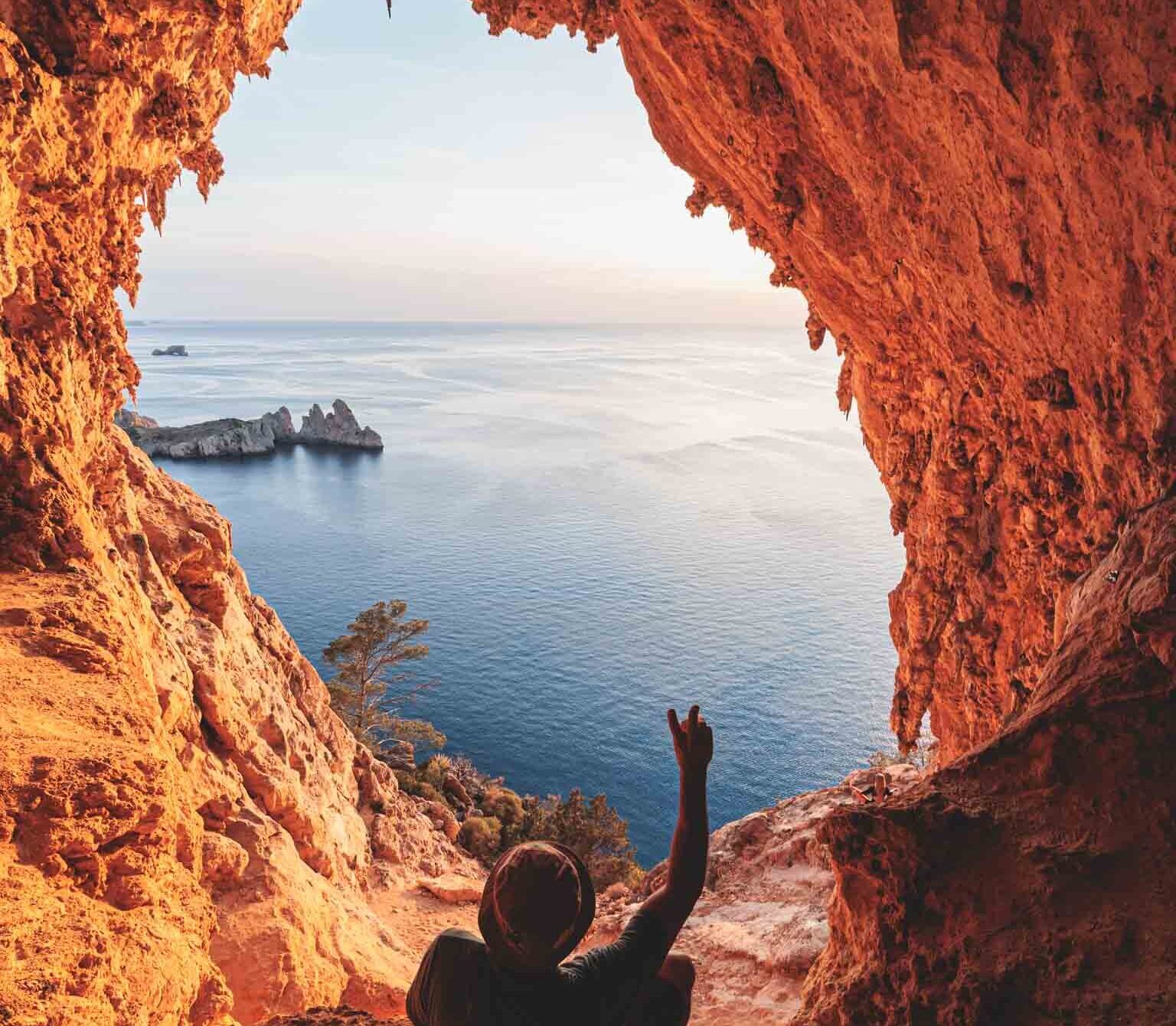 Hiker standing at the edge of a cave balcony, gazing out over sweeping views of forested cliffs and the turquoise sea on Ibiza’s remote western shoreline.