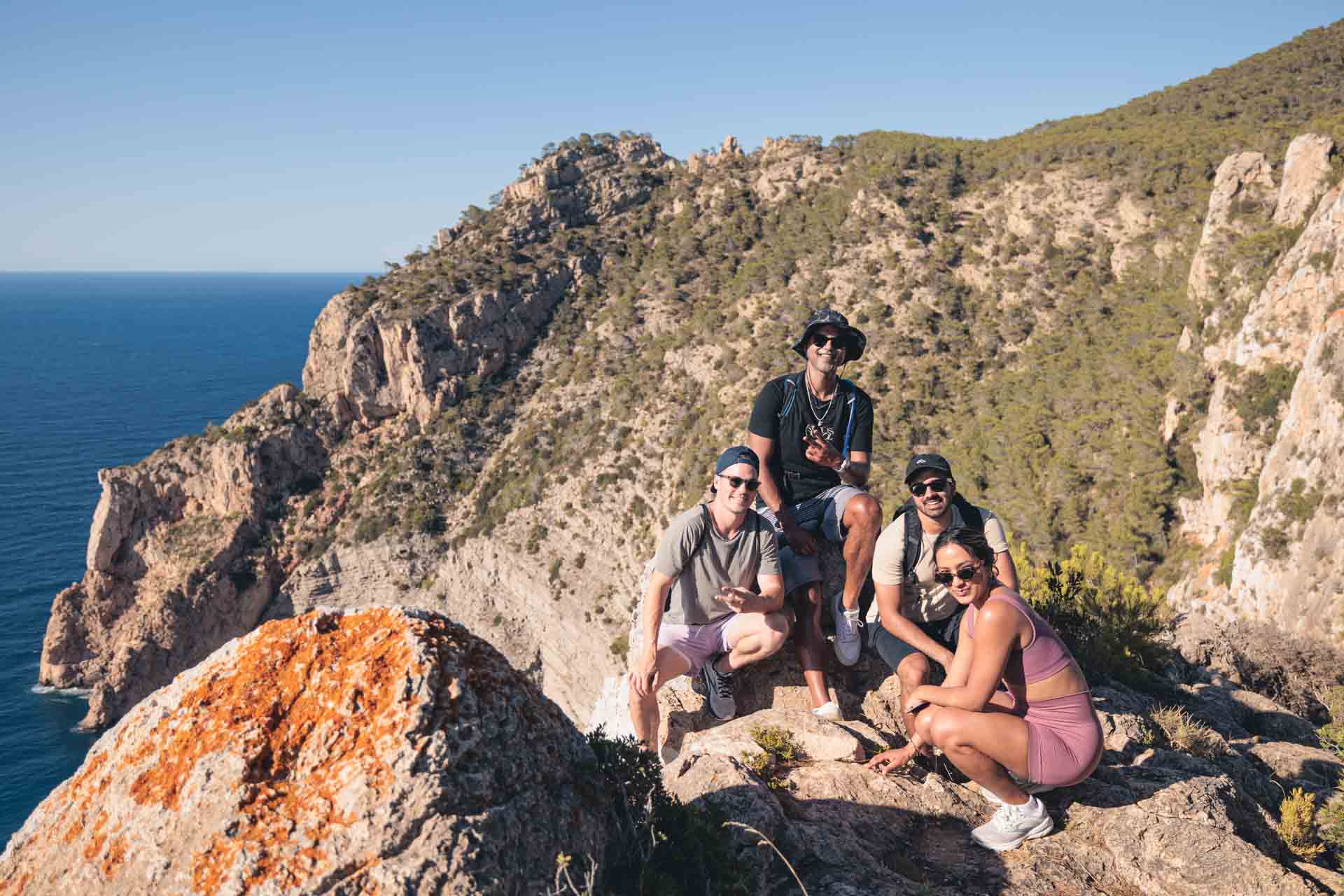Hikers navigating a steep forest trail in Ibiza, using wooden stairs and fixed ropes to descend through dense Mediterranean vegetation toward a hidden cove.