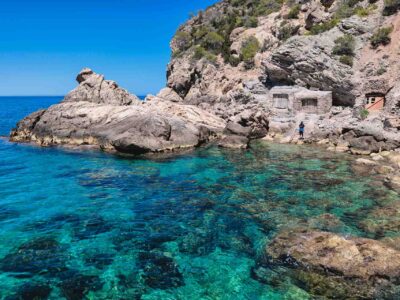 Fisherman’s Cove in Ibiza, a secluded pebble beach framed by turquoise waters, rustic fishermen’s huts, and steep cliffs, reachable only by hiking through remote coastal terrain.