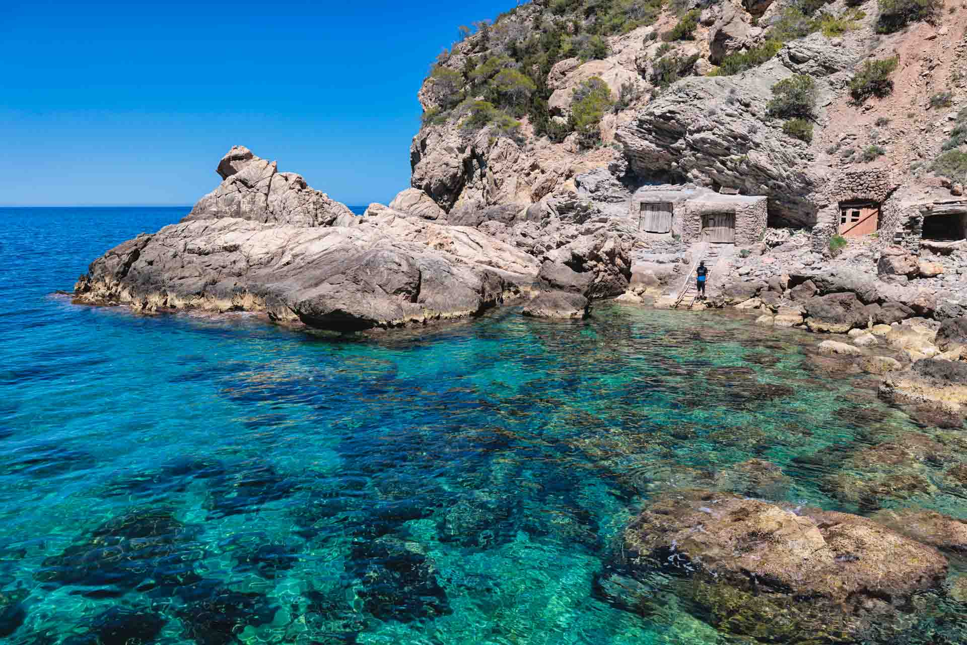 Fisherman’s Cove in Ibiza, a secluded pebble beach framed by turquoise waters, rustic fishermen’s huts, and steep cliffs, reachable only by hiking through remote coastal terrain.